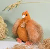 A plush hen toy sits on a surface with straw-like material and dried grass stalks. Produktbild-Vorschau