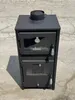 A black wood-burning stove with a chimney pipe on top, placed outdoors on a paved surface. Produktbild-Vorschau