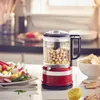 A red KitchenAid food processor filled with chickpeas is on a kitchen counter, with a plate of vegetables and a bowl of chips in the foreground. Produktbild-Vorschau