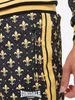 Close-up of a person's leg wearing black track pants with a gold fleur-de-lis pattern and gold stripes on the side. Náhled obrázku produktu