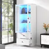 A white display cabinet with glass doors and LED lighting, placed in a room with a large window and a side table. Produktbild-Vorschau