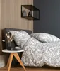 A bed with patterned bedding and pillows, next to a small wooden side table with a vase of dried flowers and a laptop. Produktbild-Vorschau