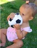 A baby sitting on grass and holding a plush soccer ball. Produktbild-Vorschau