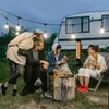 A group of people and a dog are gathered around a campfire at night, with a caravan in the background and string lights overhead. Anteprima dell’immagine prodotto