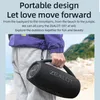 A person holding a black portable speaker on a beach with mountains in the background. Náhled obrázku produktu