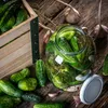 A rustic scene of cucumbers being prepared for pickling, with a wooden crate, a glass jar filled with cucumbers and dill, and garlic cloves scattered on a wooden surface. Podgląd zdjęcia produktu