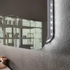 A close-up view of a modern mirror with integrated LED lighting, mounted on a textured wall. The reflection shows shelves with various items. Produktbild-Vorschau