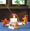A handmade candle holder with two felt figures on a seesaw, with a lit candle. Anteprima dell’immagine prodotto