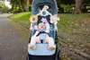 A baby is sitting in a stroller with toys attached to it, outdoors on a paved path. Produktbild-Vorschau