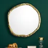 A gold-colored, irregularly shaped mirror hangs on a dark green wall above a shelf with decorative items. Podgląd zdjęcia produktu