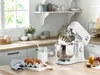 A white Kenwood stand mixer is shown on a kitchen counter, with ingredients like eggs and sugar nearby, suggesting it's being used for baking. Produktbild-Vorschau