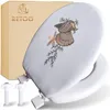 A white toilet seat cover with embroidered seashells and greenery, shown with its packaging and mounting hardware. Podgląd zdjęcia produktu