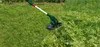 A cordless grass trimmer is being used to cut tall grass and weeds in a garden. Produktbild-Vorschau