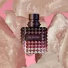 A bottle of Valentino Donna Born in Roma Intense Eau de Parfum for women is displayed prominently in the center of the image, surrounded by large, rough-hewn pinkish-white rocks against a vibrant pink background. Voir les images du produit