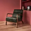 A dark green velvet armchair with wooden legs is placed in a room with pink walls and a wooden floor. A bookshelf with decorative items is visible in the background. Produktbild-Vorschau