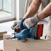 A person wearing work gloves is using a blue and red Bosch cordless planer on a wooden surface, with a window and door frame in the background. Podgląd zdjęcia produktu