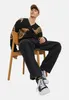 A man wearing a black cardigan with tan detailing and dark wash jeans sits on a wooden chair against a white background. Produktbild-Vorschau