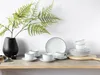 A set of white porcelain coffee cups and saucers, along with plates, are arranged on a wooden shelf, with a black vase and a fern branch in the background. Náhľad obrázku produktu