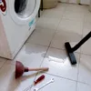 A washing machine is leaking water onto the tiled floor. A plunger, screwdriver, and wrench are on the floor near the leak, suggesting a repair is underway. A vacuum cleaner nozzle is also visible, possibly being used to clean up the water. Náhľad obrázku produktu