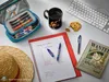 A desk scene with study and leisure items, including pens, a notebook, a mug, a cookie, a keyboard, a straw hat, and a One Piece themed book. Produktbild-Vorschau