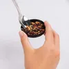 A hand is holding a small black container filled with loose tea leaves and a spoon is scooping some out. Produktbild-Vorschau