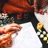 Hands painting a "paint by numbers" kit with various paint pots and knitted fabrics in the background. Produktbild-Vorschau