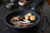 A close-up shot of a frying pan with lamb chops and potatoes, set on a wooden table with some ingredients in bowls in the background. Produktbild-Vorschau