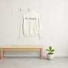 A cream-colored jacket hangs on two hooks on a white wall, with a wooden bench and a potted plant below. The floor is tiled. Produktbild-Vorschau
