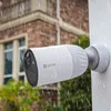 A white security camera is mounted on a white pole, with rain falling. A brick building and green foliage are visible in the background. Produktbild-Vorschau