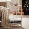 A small white side table with two mugs and a bowl of cookies on top, and books on the shelf below. A cream blanket is draped over a nearby armchair. A decorated Christmas tree and gift-wrapped boxes are in the background. Produktbild-Vorschau