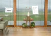 A wooden plant stand with plants and candles is placed in front of a large window with blinds. Produktbild-Vorschau