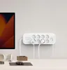 A white power strip with multiple outlets and plugs is mounted on a white wall above a desk. A portion of a computer monitor is visible on the left. Náhled obrázku produktu