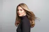 A woman with blonde hair styled in waves, wearing a black turtleneck, is posing for a photo shoot against a light gray background. Her hair is blowing to the side, creating a dynamic effect. Náhled obrázku produktu