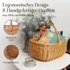 A wicker basket filled with magazines and a candle on a table, with decorative plants in the background. Produktbild-Vorschau