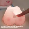 A close-up of a pink silicone night light shaped like a pig's rear end, with a finger pressing on it. Podgląd zdjęcia produktu