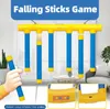 A toy falling sticks game is displayed against a window with curtains. Inset images show close-ups of the game's control panel and a side view. Náhľad obrázku produktu