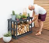 A man is placing firewood into a dark gray wooden firewood rack on a wooden deck next to a white wall. Podgląd zdjęcia produktu