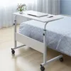 A white adjustable height table with wheels, placed next to a bed with patterned bedding. A laptop and a mouse are on the table, along with a small vase of flowers. Voir les images du produit