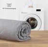 A rolled-up gray blanket is placed on a wooden surface in front of a washing machine. Produktbild-Vorschau