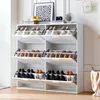 A white shoe cabinet with multiple shelves and flip-down doors, filled with shoes, placed in a room with a framed picture and a camera on top. Anteprima dell’immagine prodotto