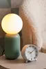 A green lamp and a white alarm clock sit on a table next to a textured chair. Anteprima dell’immagine prodotto