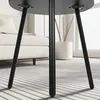 Close-up of the legs of a black round table on a striped rug, with a white sofa and a potted plant in the background. Produktbild-Vorschau