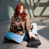 A young woman with red hair sits on the ground in an urban setting, holding a smartphone. Náhľad obrázku produktu