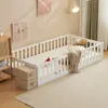 A white wooden children's bed with railings and a mattress, set up in a room with artwork on the wall and a floor lamp. Náhľad obrázku produktu