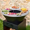 A grill-fire pit with food cooking on it, set against a stone wall and grass. Anteprima dell’immagine prodotto
