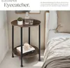 A round, two-tiered side table with a wood-grain finish and black metal legs is placed next to a bed with white and beige bedding. A small potted plant, a glass bottle, and a glass are on the top shelf, while a book and a decorative object are on the botto Náhled obrázku produktu