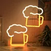 Two beer mug-shaped neon lights are displayed on a table, one in the foreground and one slightly behind and to the right. A small decorative Christmas tree is visible on the right side of the image. Náhled obrázku produktu