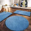 Three blue bath mats are arranged on a wooden floor in a bathroom setting, with a stool, basket, and artwork visible in the background. Anteprima dell’immagine prodotto