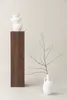 A minimalist studio shot featuring two white vases and a wooden pedestal against a light gray background. Produktbild-Vorschau
