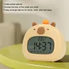 A cute, cartoonish alarm clock shaped like a capybara is placed on a green table. The clock displays the time and temperature. Náhled obrázku produktu